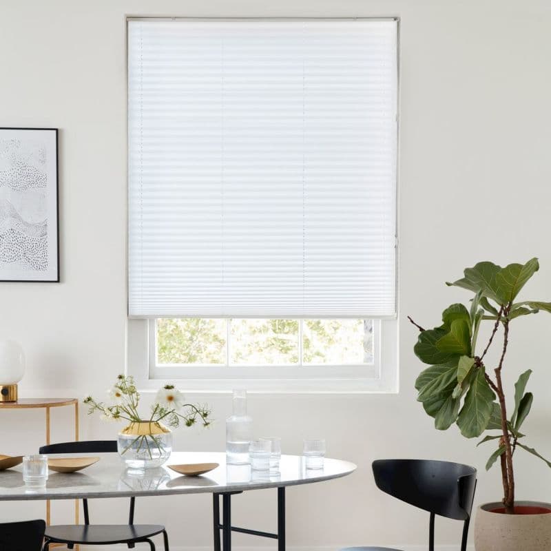 White pleated cellular blind, lowered nearly to the sill with horizontal seams, diffusing daylight; set in a white dining area with marble table, black chairs and a potted fiddle‑leaf fig.