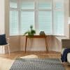 Horizontal white Venetian blinds cover a five-panel bay window, closed and filtering daylight; wooden console holds potted plants in a living room with wood floor, rug, chair and sofa.