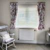 Pink floral pleated curtains, tied back on a silver rod, frame a window; white slatted blinds filter daylight into a pale room with a radiator and a cushioned rocking chair.