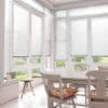 White horizontal venetian blinds cover tall bay windows, lowered and filtering soft daylight; overlooking a green garden, bright dining area with a white table, wooden chairs, and floral cushions.