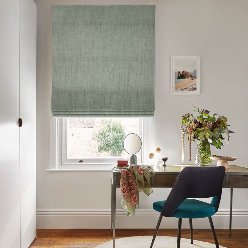 Sage-green fabric Roman blind, lowered with a folded hem; filtering daylight through a lower gap over a desk in a bright home office with a teal chair and vase of flowers.
