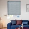 Light-gray pleated cellular blind lowered over the window, filtering daylight through horizontal ribs; above a blue sofa in a modern living room with cushions, floor lamp, and partial roof-and-tree view.