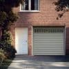 Sheer white curtains (upper window) — closed, softly diffusing daylight — against a red brick house façade with a white side door, closed roll-up garage door, driveway and leafy shrubs.