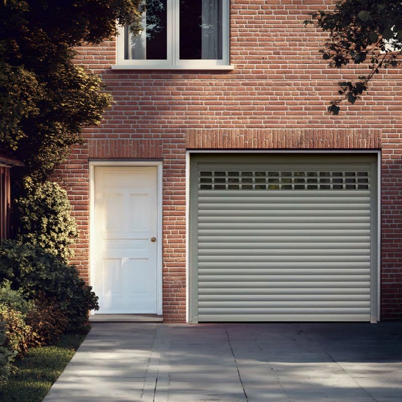 Sheer white curtains (upper window) — closed, softly diffusing daylight — against a red brick house façade with a white side door, closed roll-up garage door, driveway and leafy shrubs.