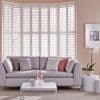 White full-height plantation shutters across a multi-panel bay window, closed with slats filtering bright daylight; behind a pale gray sofa with cushions, coffee table, orchid, and soft pink walls.