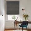Dark grey Roman shade, fully lowered with subtle folds at the bottom, filtering daylight through a white sash window; above a wooden desk with teal chair, vase of flowers and a small mirror.