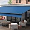 Blue retractable fabric awning with black front bar, fully extended over sliding glass doors, shading a modern outdoor patio with wicker chairs, low wooden table, brick wall and daylight.