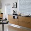 Horizontal Venetian blinds covering a wide window and door, fully closed and filtering daylight; bright modern kitchen with light wood cabinets, black countertop, bar stools and glass jars.
