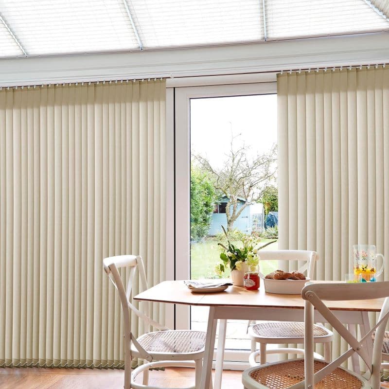 Cream vertical blinds, floor-to-ceiling on a sliding glass door, parted at center with slats mostly closed, filtering daylight into a bright dining nook with wooden table, chairs, and garden view.