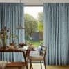 Floor-length patterned curtains (teal geometric fabric) are pulled aside, framing a central glass door and admitting soft daylight; a dining table and chairs set with breakfast face a leafy backyard.