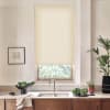 A beige roller blind covers a kitchen window, filtering daylight over a countertop with a stainless sink and faucet, bowls of leafy greens, potted plants, and framed wall art.