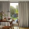 Floor-length, pinch-pleat grey curtains, drawn open at the center and hanging straight, framing a sunlit backyard; a wooden dining table set for breakfast with pancakes and a pendant light in foreground.