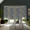 Patterned roller blinds cover French glass doors, partially lowered, in a modern living room with green armchairs, a glass coffee table on a chevron rug, and a garden visible outside.