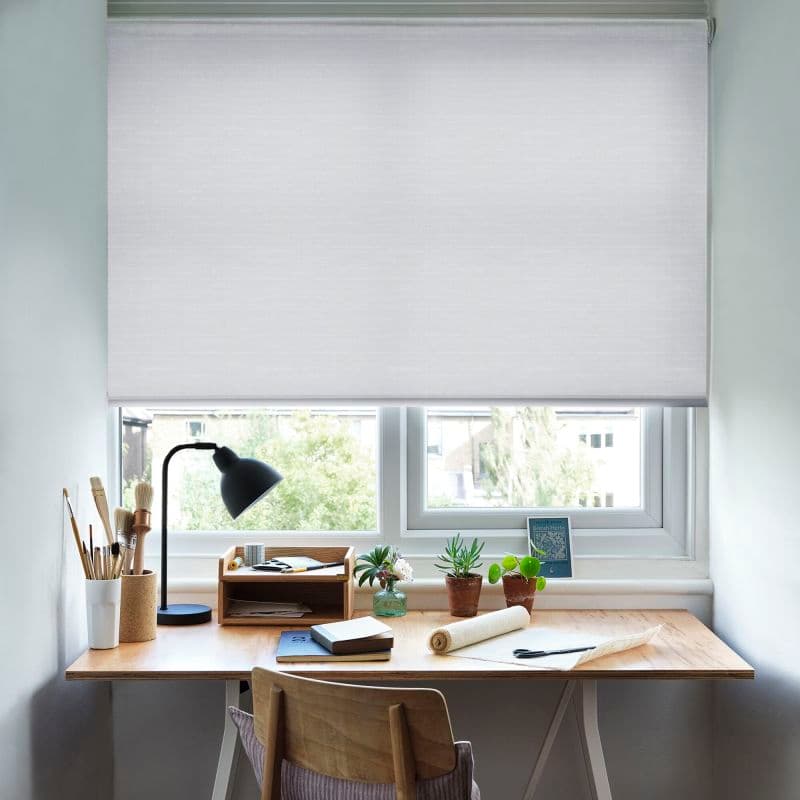 White roller blind, lowered across the window, semi-opaque fabric filtering soft daylight; desk beneath holds a lamp, art brushes, notebooks and potted plants in a bright, tidy home workspace.