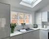 Horizontal white Venetian blinds covering a three-panel window, lowered with slats slightly open, filtering daylight over a modern white kitchen sink filled with soapy water; skylight overhead, fenced yard view.