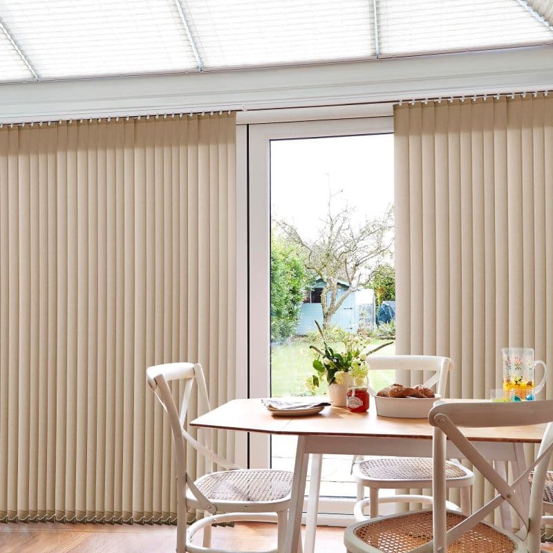 Beige vertical blinds cover sliding glass door, panels mostly closed with a central gap revealing view; filtering daylight into a dining area with wooden table, white chairs, and garden beyond.