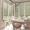 Green Venetian blinds on tall windows and patio doors, lowered and filtering daylight while leaving lower panes visible; bright sunroom dining area with white table, chairs, cushions, garden view.
