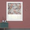Floral-patterned Roman shade, partially lowered with visible soft folds, diffusing daylight; installed in a white-trimmed window set in a mauve wall, above a teal dresser and gold circular wall sconce.