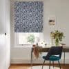 Navy-and-white leaf-pattern Roman blind lowered over a sash window, partially blocking light; small desk beneath with mirror, vase of flowers and teal chair in a bright, neutral study.