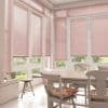 Pink horizontal Venetian blinds lowered over tall paneled windows and French doors, slats mostly closed, filtering daylight into a bright dining nook with wooden table, chairs and garden glimpses.
