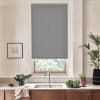 Grey patterned roller blind hangs, partially lowered over a window above a sink, in a bright kitchen with faucet, wooden cabinets, fresh greens and potted plants.