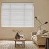 White horizontal blinds covering a rectangular window, closed and filtering soft daylight, in a beige living room with a sofa, wooden coffee table holding a vase and open book.