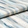 Low-pile woven rug with blue, gray, and cream abstract streaks; folded over itself; tight close-up studio view emphasizing woven texture and layered edges.