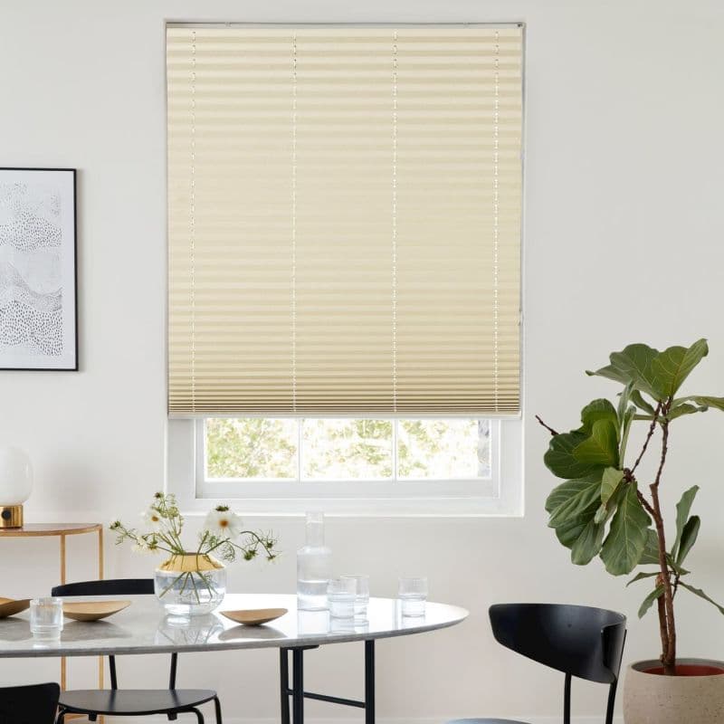 Beige pleated blind lowered over a window, closed and gently filtering daylight through horizontal folds; set above a dining table with glassware, chairs, potted fiddle‑leaf fig and minimalist wall art.