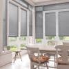 Grey horizontal Venetian blinds lowered over tall French doors and surrounding windows, filtering soft daylight into a bright dining nook with white wooden table, chairs and a visible garden view.