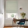 Roman blind with pale green-and-cream geometric pattern, lowered over the window, filtering soft daylight into a home office with desk, teal chair, vase of foliage, mirror and coastal print.