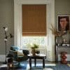 Brown patterned Roman blind (diamond motif) lowered over a white-framed sash window, filtering daylight; set in a cozy living room with green armchair, low carved table, lamp, plants, framed portrait.