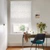 A white Roman blind patterned with small multicoloured stars hangs down in neat folds, filtering soft daylight over a desk with mirror, chair and vase of flowers in a neutral study.