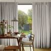 Full-length grey pleated curtains pulled aside to frame a sliding glass door, filtering soft daylight; wooden dining table set for breakfast with pancakes, a pendant lamp, and a green garden beyond.