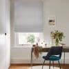 Grey patterned Roman blind lowered over window, filtering soft daylight; a wooden desk with mirror, vase of flowers and draped scarf; bright home office with neutral walls and teal chair.