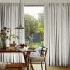 Floor-length light-gray pleated curtains, parted to reveal a sliding door, filtering soft daylight; a wooden dining table with plates and a pendant lamp faces a sunlit garden with trees.