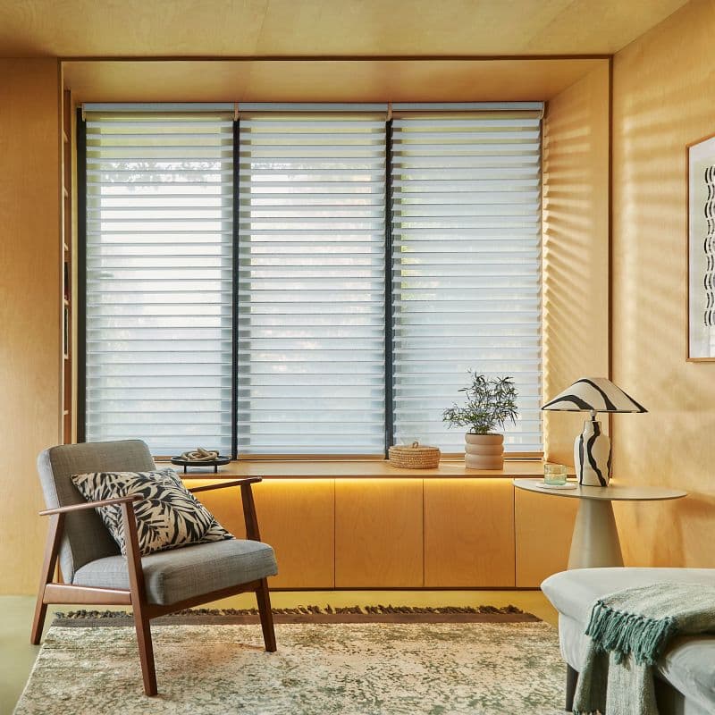 Layered horizontal sheer-and-opaque roller shades covering three tall window panels, partially closed and filtering soft daylight; set in a warm plywood-walled sitting nook with armchair, side table, lamp, and plant.
