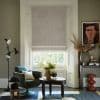 Pale beige fabric Roman blind lowered over a sash window, softly filtering daylight; cozy living room setting with green swivel armchair, textured coffee table, plants, muted walls and framed portrait.