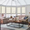 Cream honeycomb pleated blinds cover tall conservatory windows, lowered mid-height and softly diffusing daylight; cozy sunroom with beige sofa, pink knit throw, wooden coffee table and blue rug.