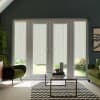 Glass patio doors filter daylight through narrow vertical striped blinds, framing a sunlit garden beyond; set in a teal-walled living room with green armchairs, glass coffee table and chevron rug.