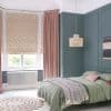 Floral-patterned roman blinds lowered over a bay window with long dusty-pink floor-length curtains drawn aside; they filter soft daylight into a teal-paneled bedroom with a green bed, woven pendant and round jute rug.