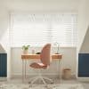 White horizontal blinds covering a wide window, slats tilted to filter bright daylight; small attic-style home office with wooden desk, pink swivel chair, desk lamp, potted plant, neutral decor.