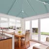 Teal pleated roof blinds cover the conservatory ceiling panels, meeting at the apex; fully lowered, diffusing light. White pleated window and door blinds closed; wood table, beige chairs, garden view.