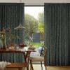 Dark green floor-length botanical-print curtains, parted to frame a glass door and filter soft daylight; dining table and pendant light in foreground, sunlit garden with trimmed trees beyond.