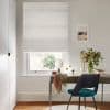 A patterned white Roman blind lowered over a sash window, softly filtering daylight. Below, a wooden desk with teal chair, mirror, vase of greenery, framed seaside print on pale wall.