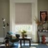 Textured beige Roman shade, lowered over a tall sash window with a narrow light gap at the bottom; filtering daylight into a cozy living room with armchair, carved coffee table, console and artwork.