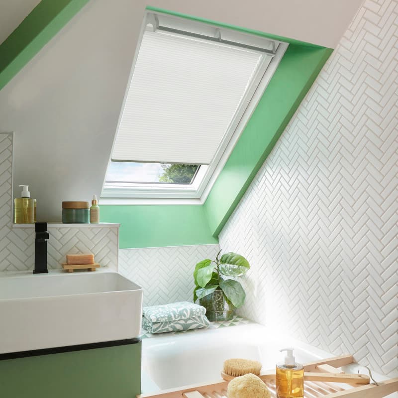 White pleated skylight blind, partially lowered and filtering soft daylight, in a sloped bathroom with mint-green window trim, white herringbone tiles, sink and bathtub with plant and bath tray.