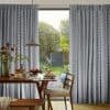 Gray patterned, floor-to-ceiling curtains, parted centrally and filtering soft daylight; a wooden dining table set with pancakes and fruit, pendant lamp above, chair and sunlit garden visible outside.