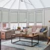 Pale cellular pleated blinds, lowered to mid-height across tall conservatory windows, filtering soft daylight; beige sofa with chunky pink throw and cushions; wooden coffee table on blue rug.