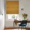 Golden-yellow Roman blind, lowered with soft folds, filtering daylight; over a white sash window above a desk with chair, vase of flowers and framed seascape in a bright neutral room.