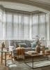 White horizontal Venetian blinds across a bay window, slats mostly closed and filtering soft daylight; in front, a light-gray sofa with cushions, wooden tables, vase, and warm neutral living room.